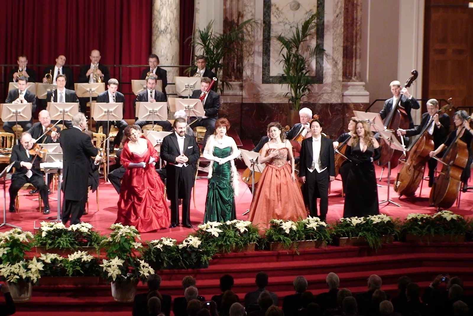 Six opera soloists in red, green, orange and black gowns and evening dress stand in front of the string players on the stage of the Hofburg Festival Hall