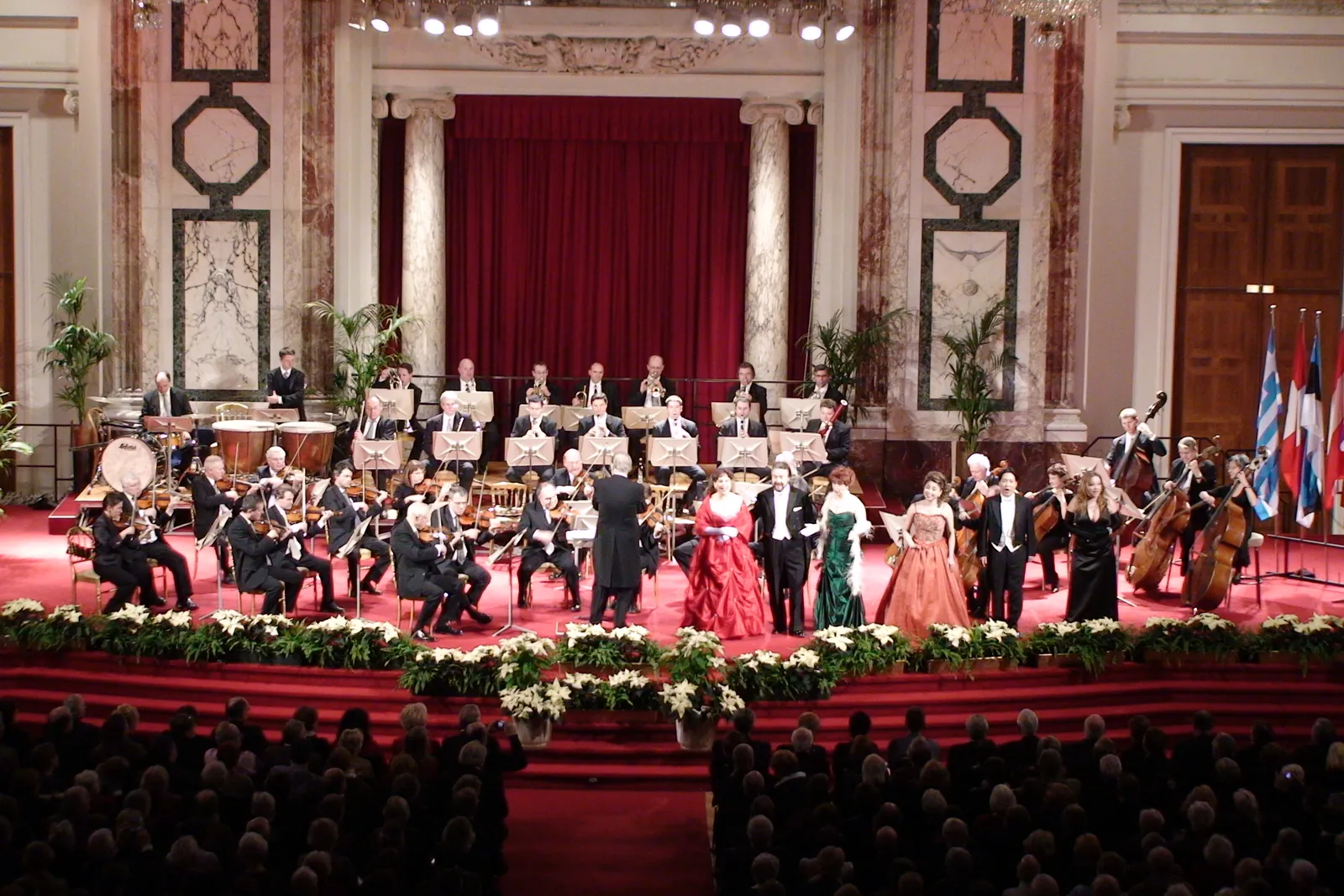 The Vienna Hofburg Orchestra and five soloists in evening dress on a red-carpeted stage in the Hofburg Festival Hall, with a crimson curtain and marble pilasters behind