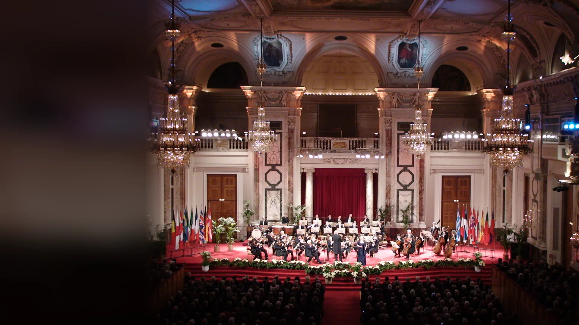 The Vienna Hofburg Orchestra on a red-carpeted stage in the Hofburg Festival Hall, under crystal chandeliers, marble pilasters and a crimson curtain, with the audience in front